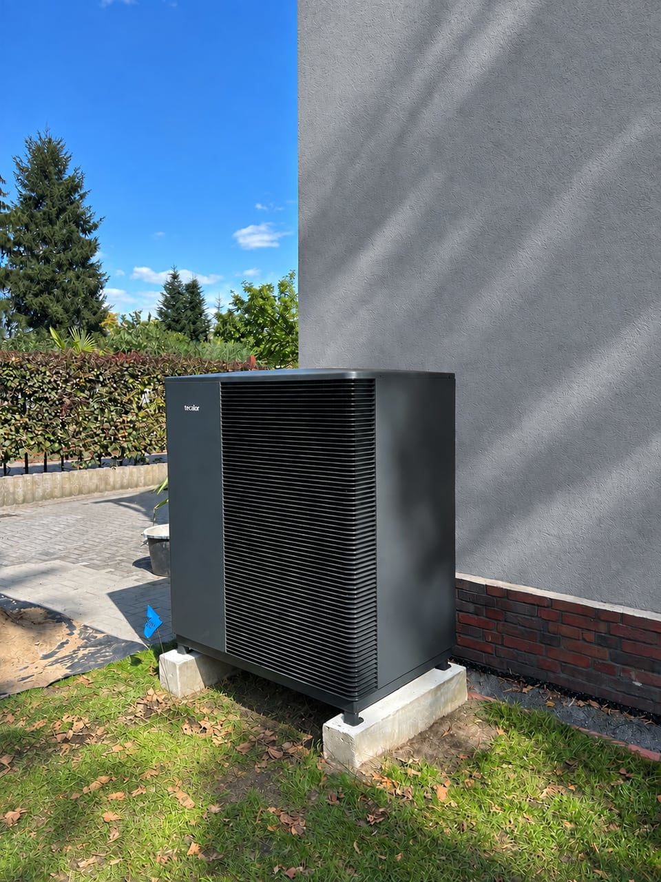 Dunkles Wärmepumpen-Außengerät auf Betonstützen neben einer Hauswand.